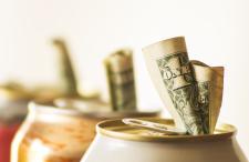 A close-up view of aluminum soda cans with money placed inside the drinking spout against a white background.